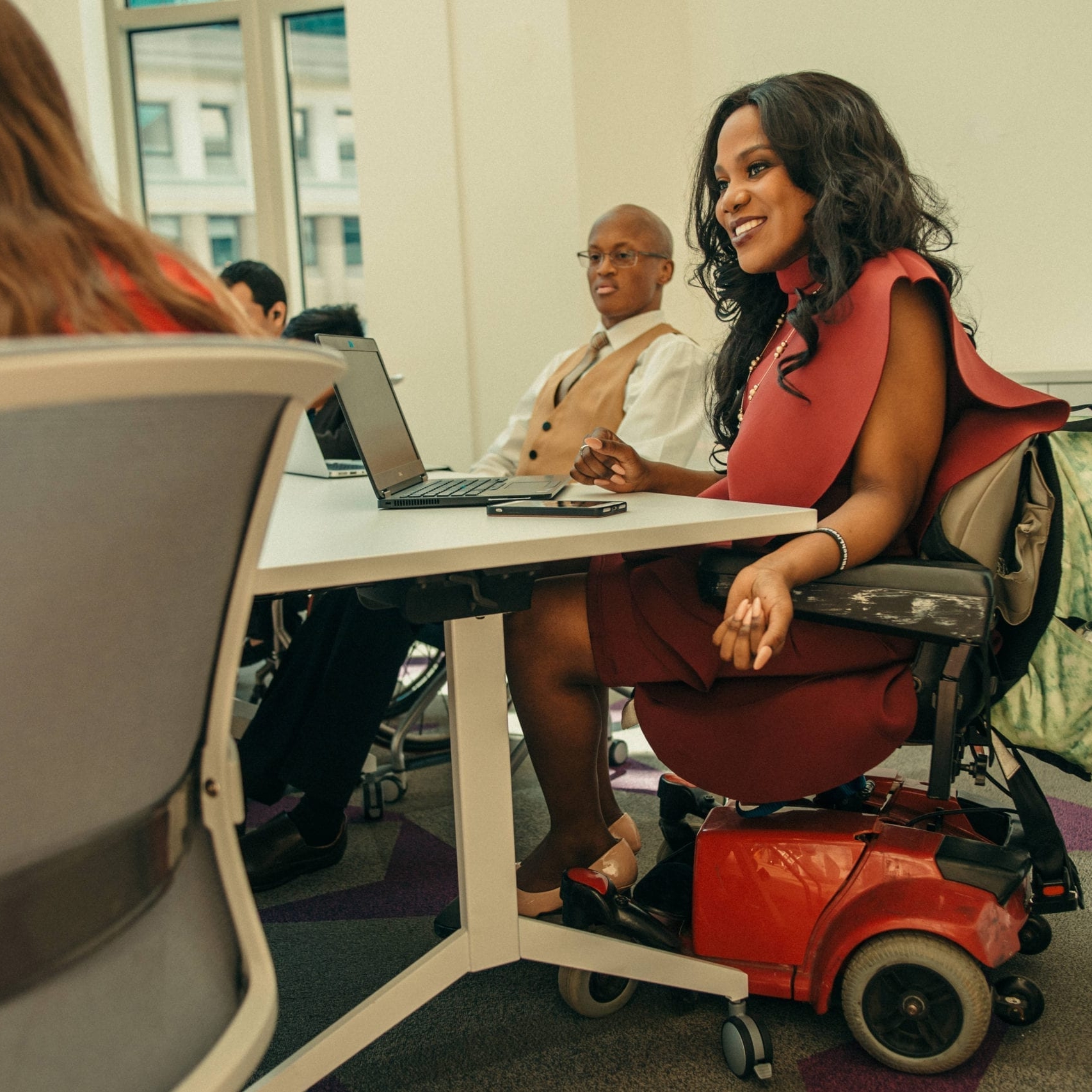 Group of people sit around a board room, focus on women dressed professionally in a motorized wheelchair.