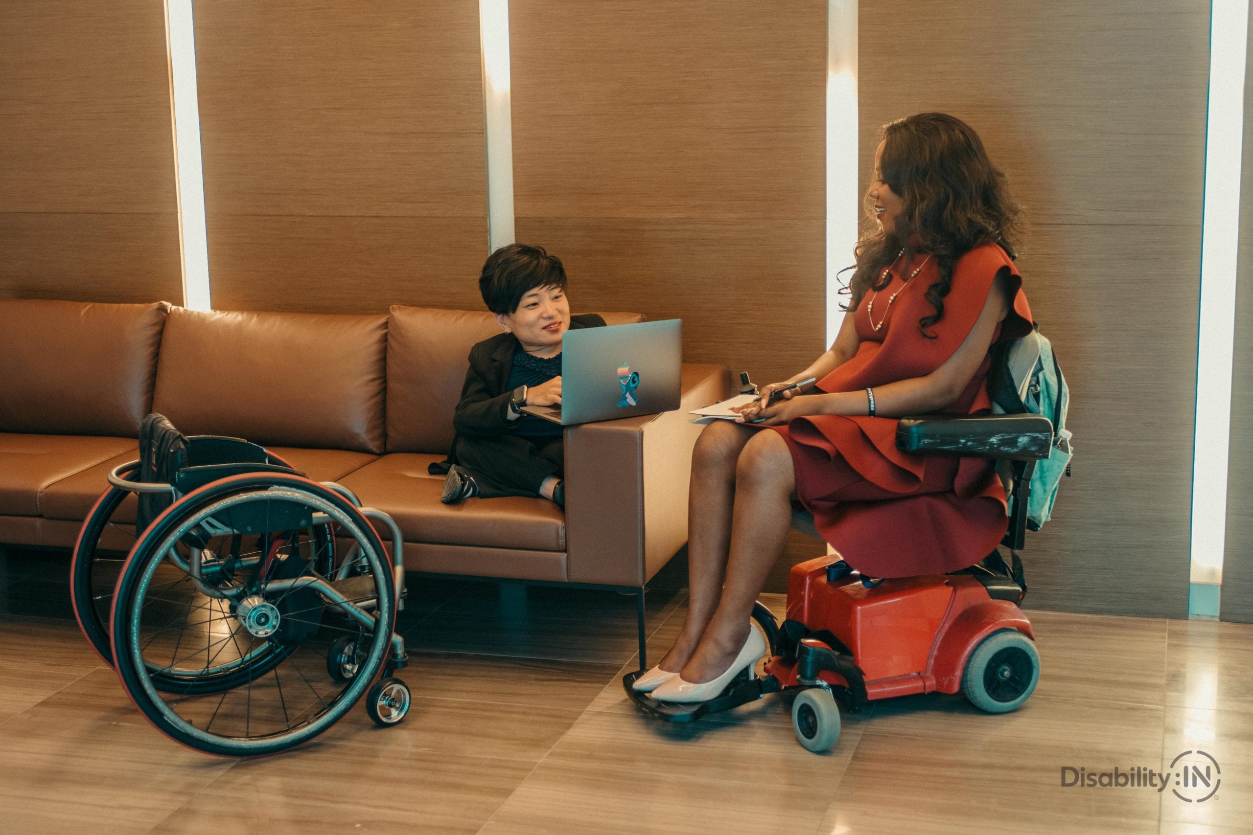 Two women in professional setting discuss work. Both women utilize a wheelchair.