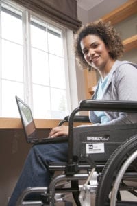 Woman in a wheelchair using a laptop and looking at camera.