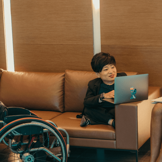 A person of short stature sitting on a couch, using a laptop, with their wheelchair next to the couch. They are wearing a black suit and have cropped black hair. They are speaking to a woman with long black hair in a flowing red dress.