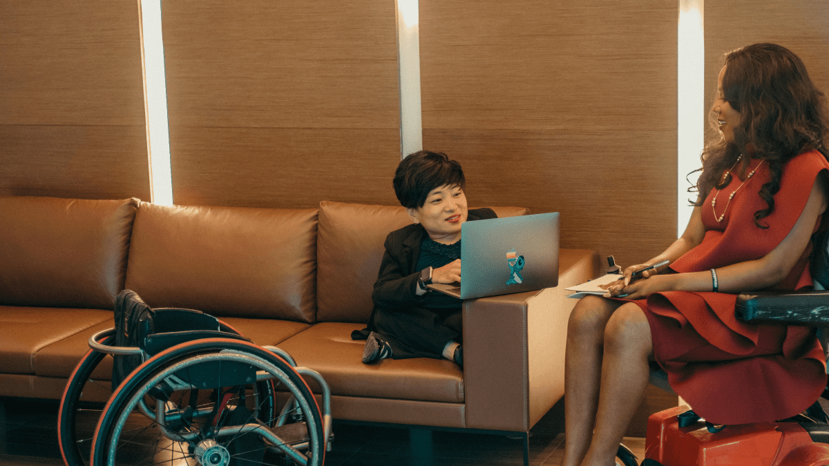A person of short stature sitting on a couch, using a laptop, with their wheelchair next to the couch. They are wearing a black suit and have cropped black hair. They are speaking to a woman with long black hair in a flowing red dress.