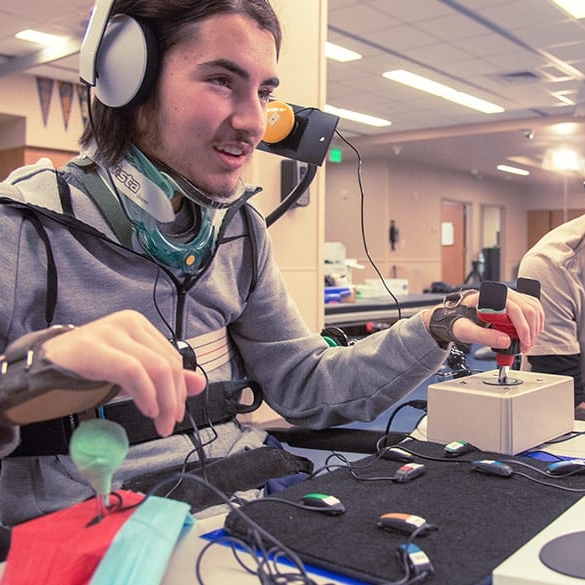 A person testing out Microsoft's new Xbox Adaptive Controller with another person nearby. The person using the controller is wearing a head set and has fair skin and is wearing a headset. The other person has black hair and is wearing glasses and is watching them with a smile.