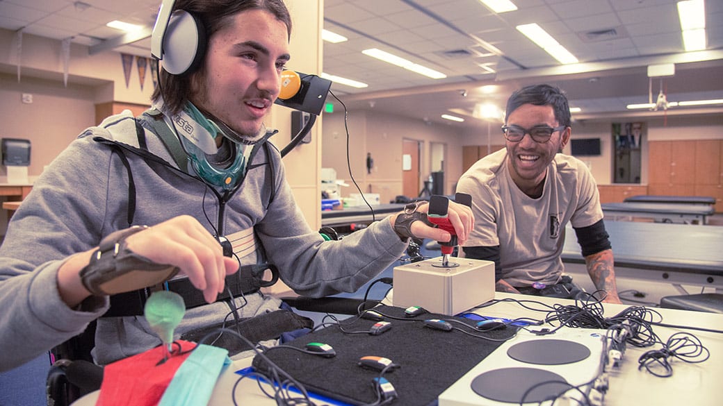 A person testing out Microsoft's new Xbox Adaptive Controller with another person nearby. The person using the controller is wearing a head set and has fair skin and is wearing a headset. The other person has black hair and is wearing glasses and is watching them with a smile.