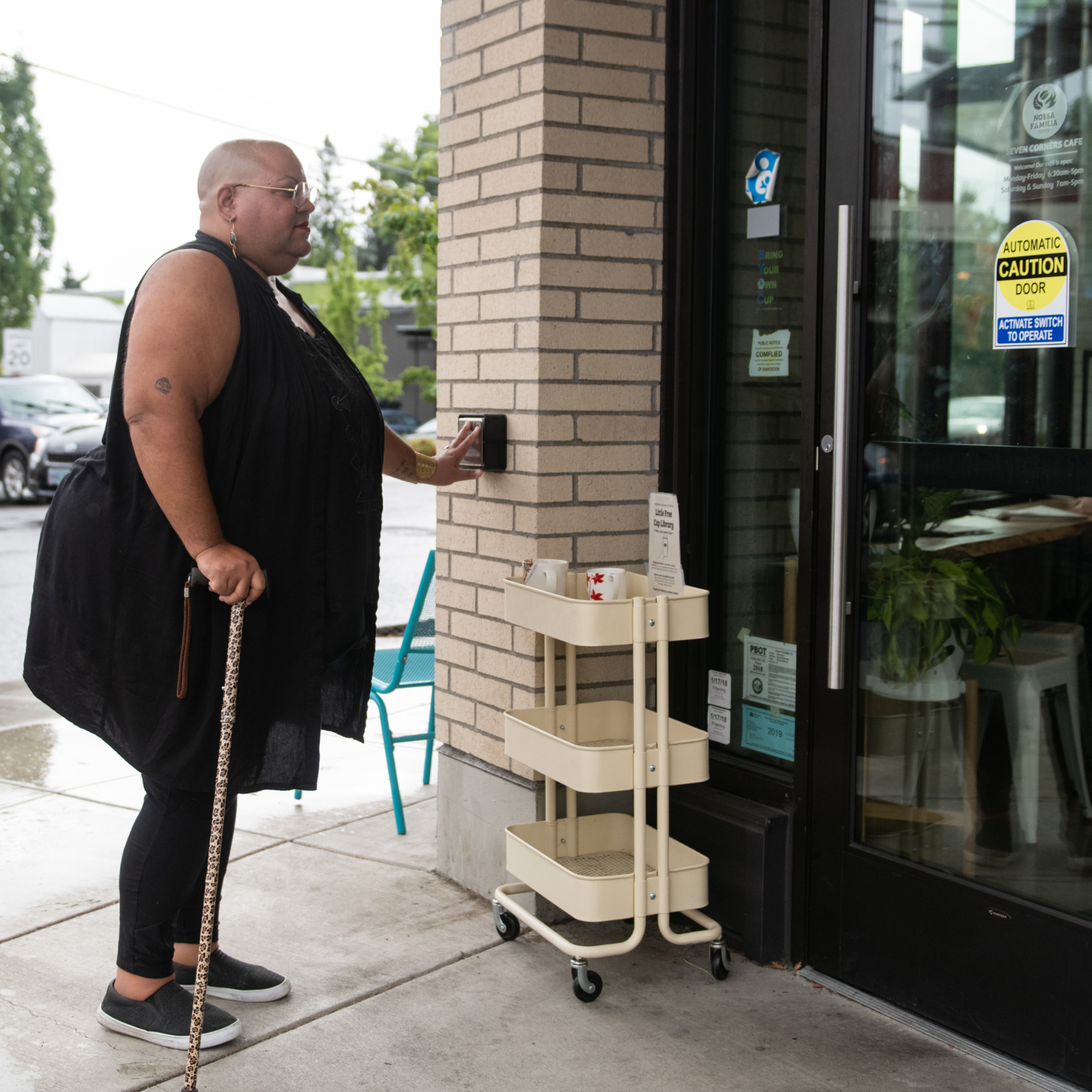 A Black non-binary person with a leopard print cane presses a wall-mounted push button to open the cafe door. They are dressed in all black and have a shaved head, glasses, and a red lip on.