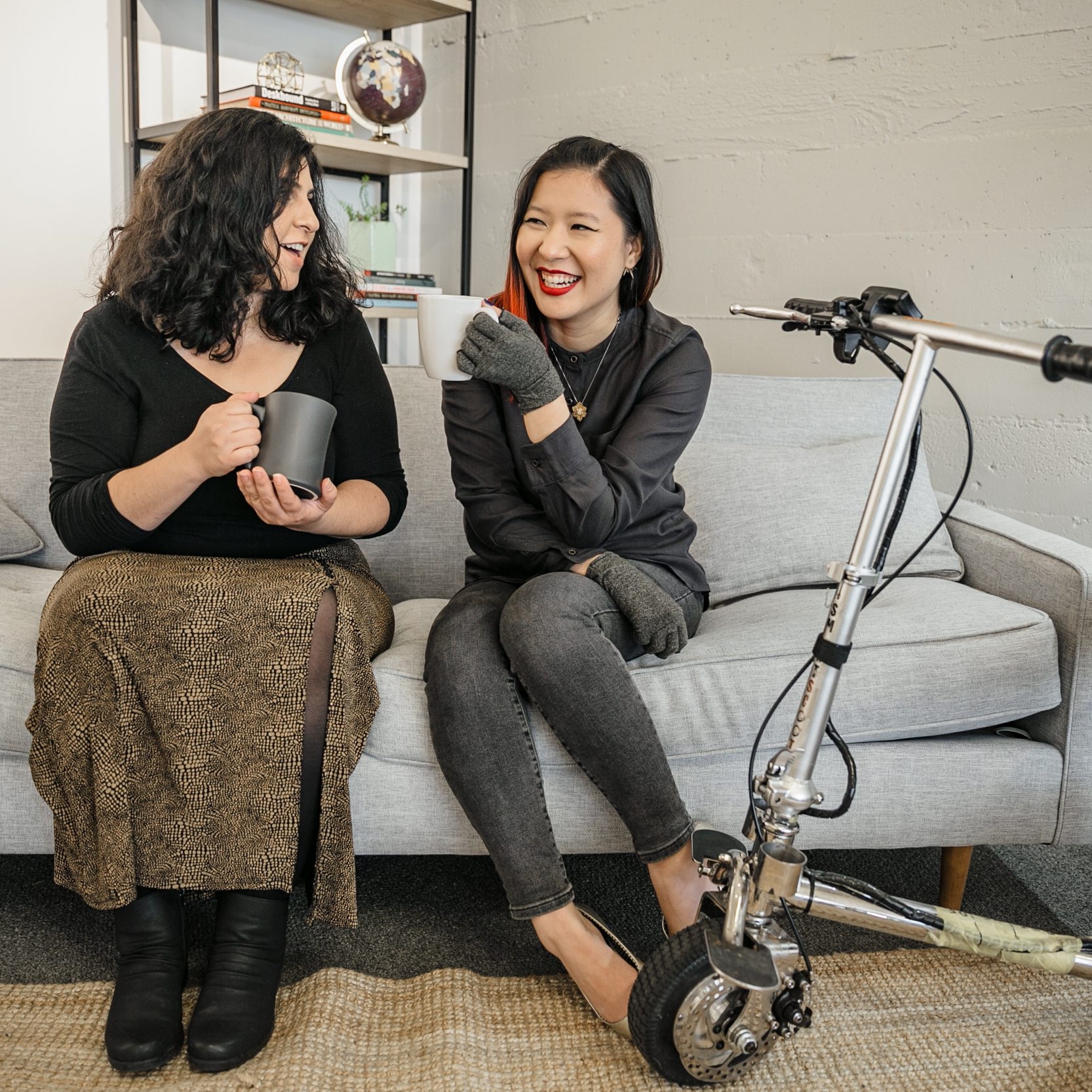 A Latinx disabled woman and an Asian disabled genderfluid person chat and sit on a couch, both holding coffee mugs. An electric lightweight mobility scooter rests on the side.