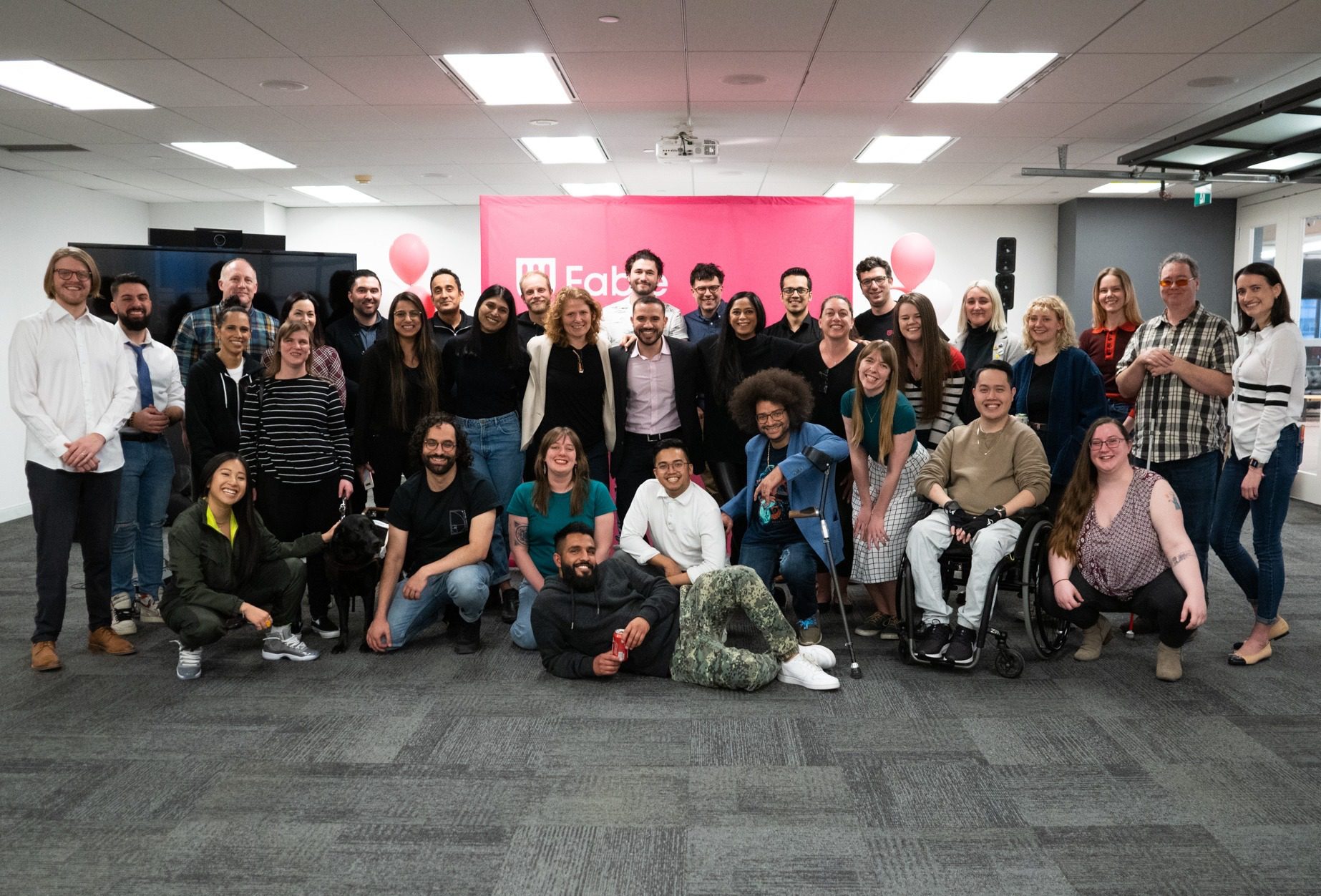 A group of 35 Fable employees standing and sitting together with big smiles. Some people sit and kneel in the front row, including a black guide dog. A pink Fable backdrop and pink and white balloons are in the background.