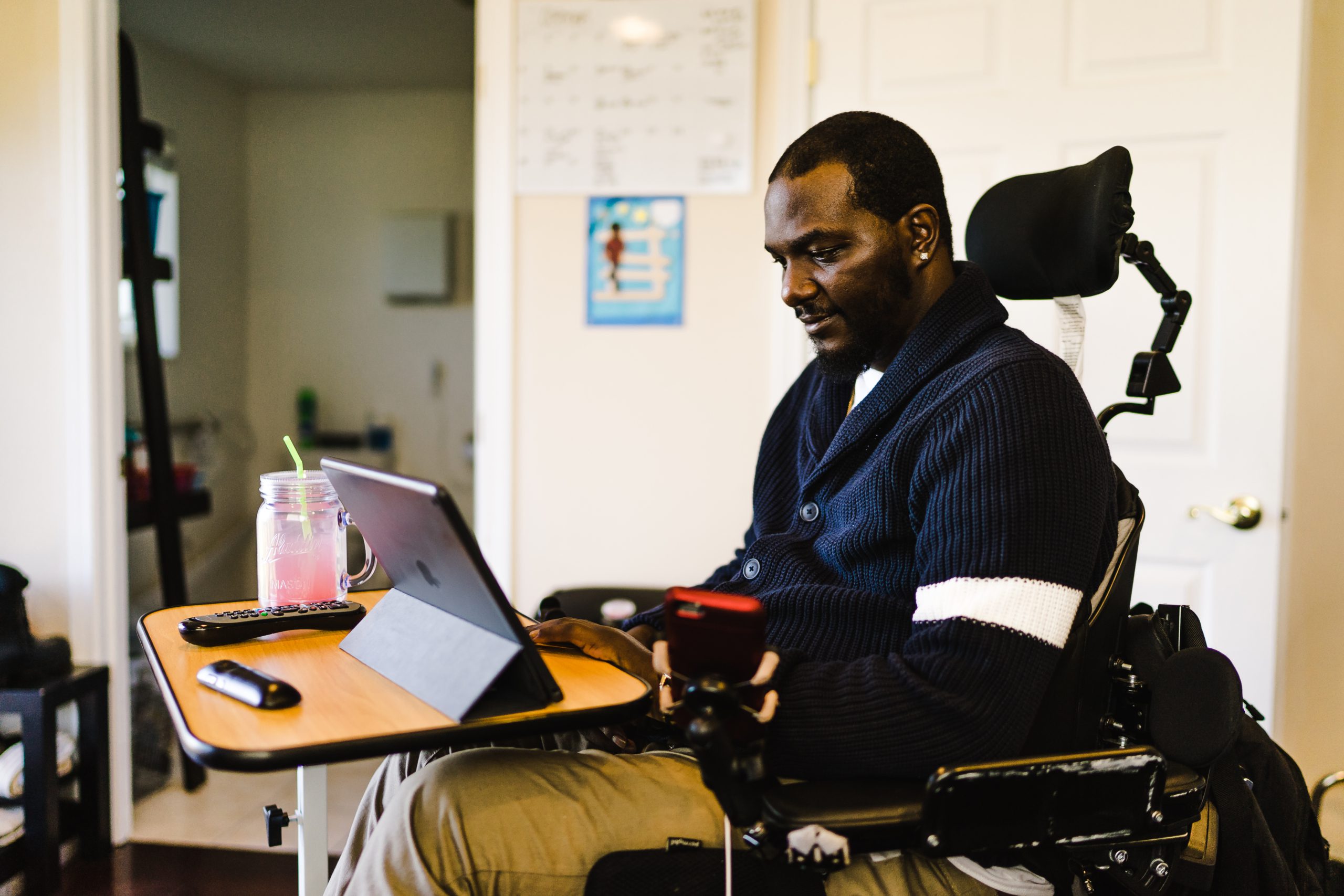 A Black man in a powered wheelchair sits at a table, watching a tablet.