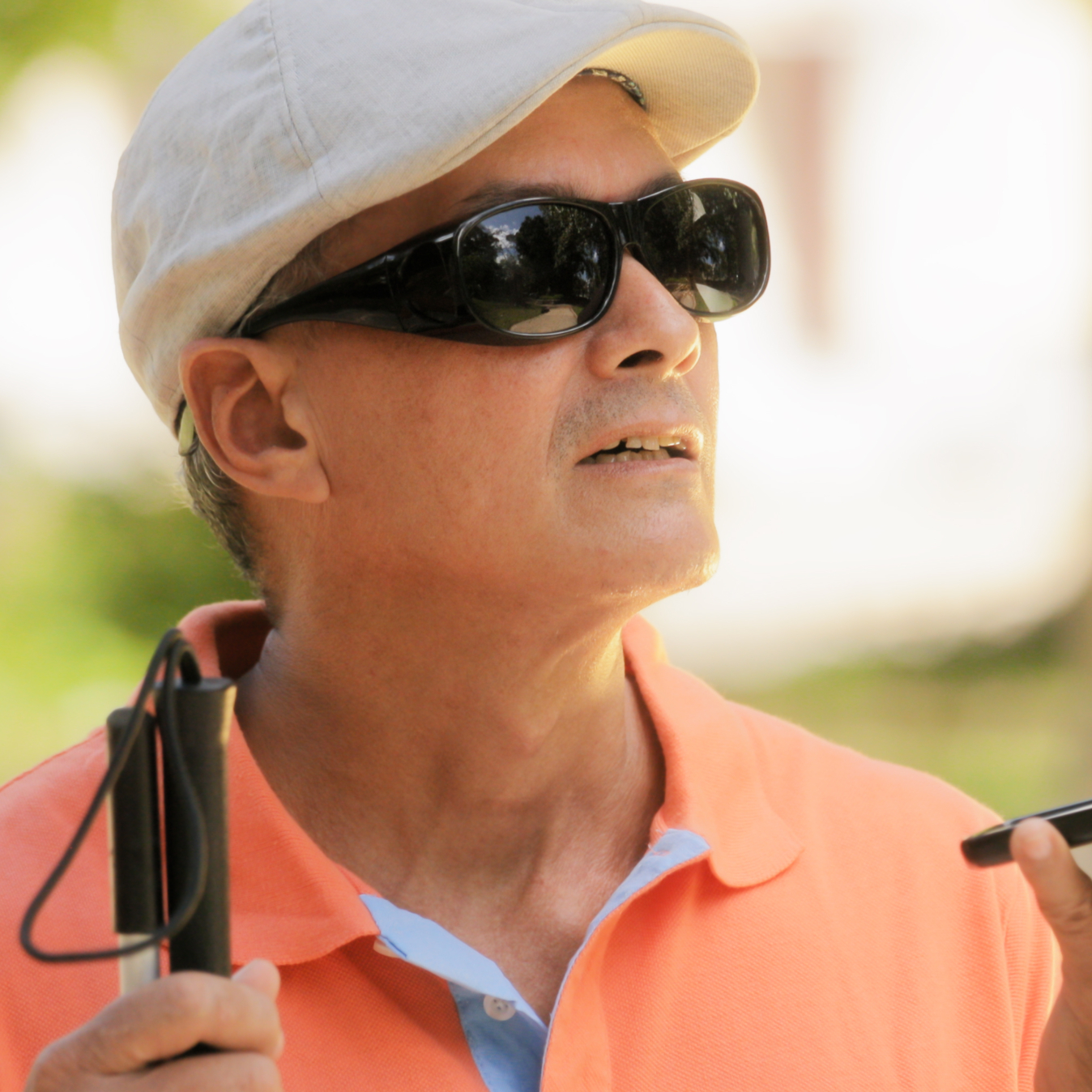 Blind man with light brown skin holds a smartphone in front of him as he stands outside. He wears opaque black sunglasses, a flat cap and it holding a white cane.
