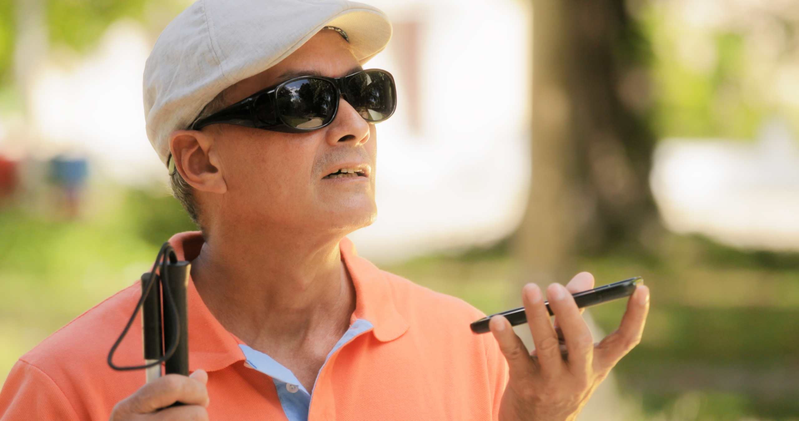 Blind man with light brown skin holds a smartphone in front of him as he stands outside. He wears opaque black sunglasses, a flat cap and it holding a white cane.