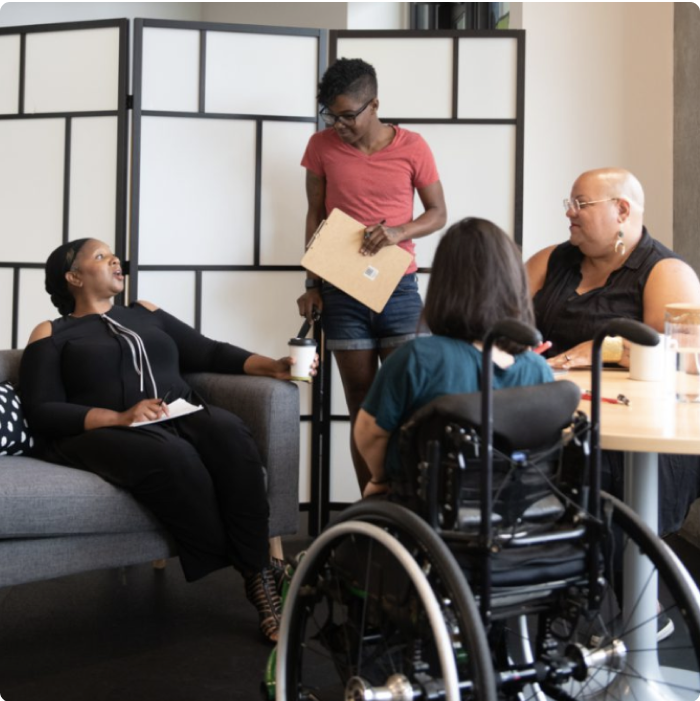 Four disabled people of color gather around a table during a meeting. A Black woman sitting on a couch gestures and speaks while the three others (a South Asian person sitting in a wheelchair, a Black non-binary person sitting in a chair, and a Black non-binary person standing with a clipboard and cane) face her and listen.