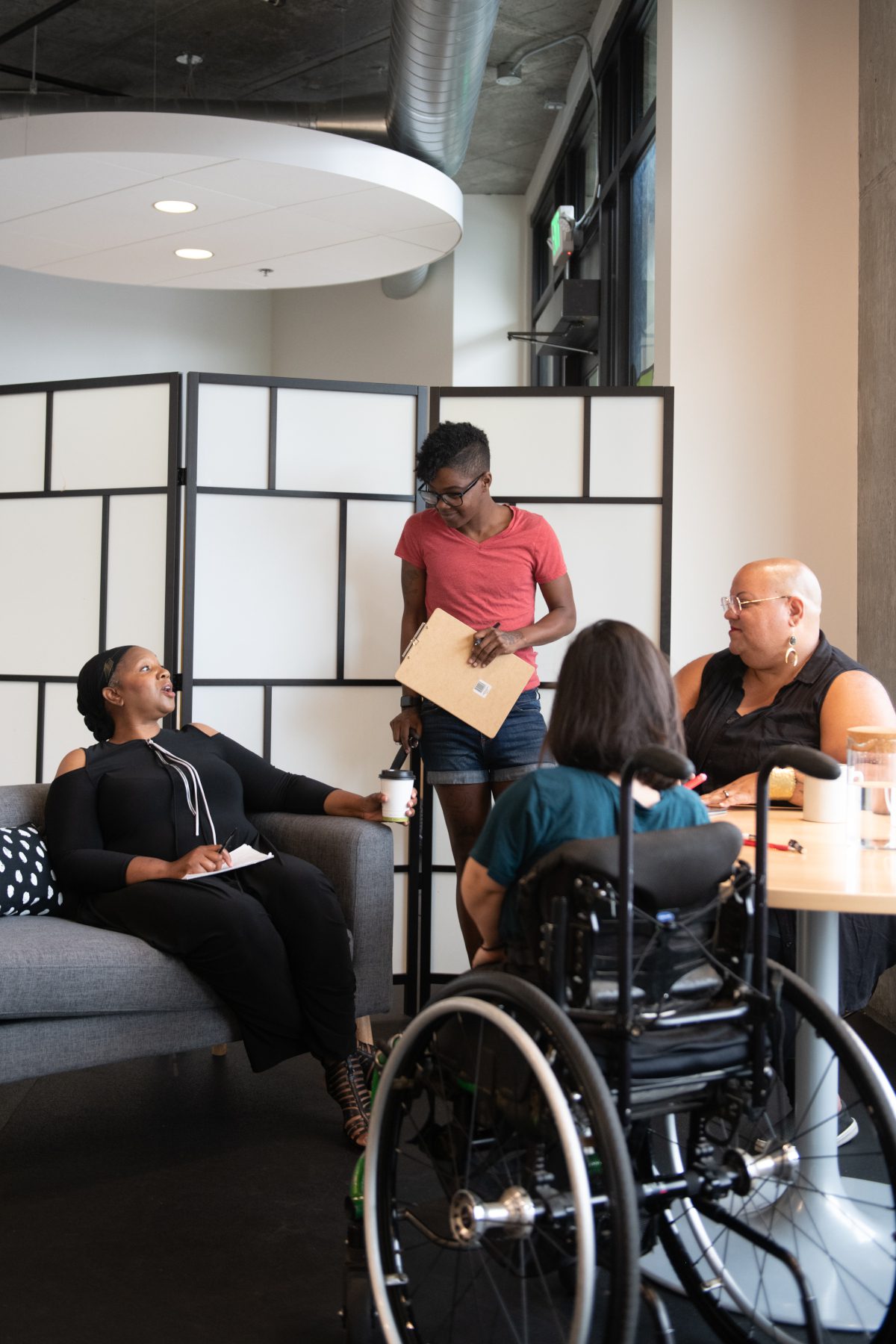 Four disabled people of color gather around a table during a meeting. A Black woman sitting on a couch gestures and speaks while the three others (a South Asian person sitting in a wheelchair, a Black non-binary person sitting in a chair, and a Black non-binary person standing with a clipboard and cane) face her and listen.