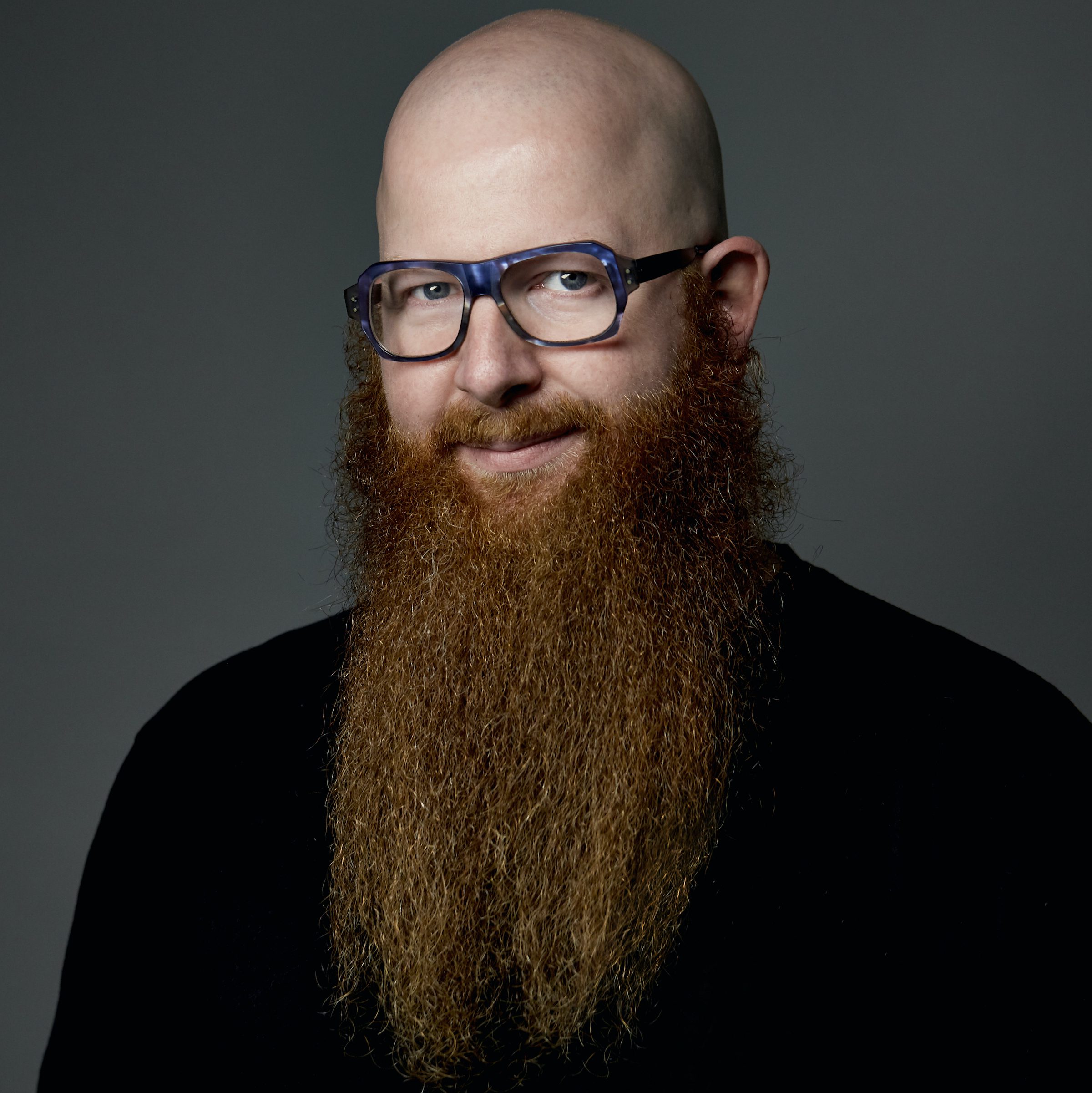 Greg is a white man wearing a black sweater. He is bald and wears a long beard.