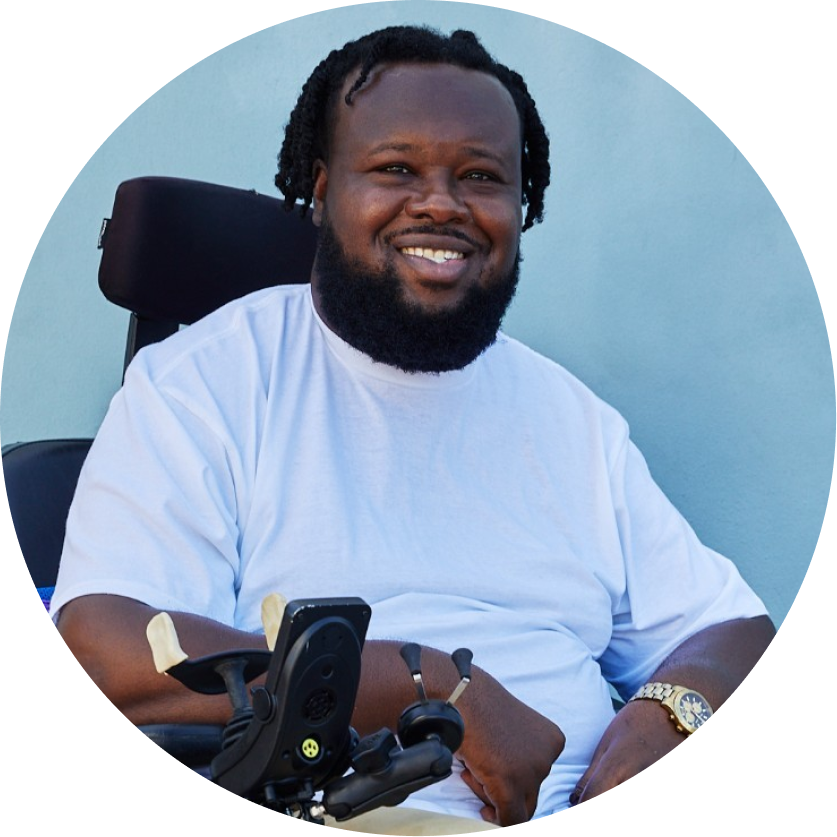 Headshot of Rhonel Cirous, a Black man with short twists smiling in a power wheelchair.