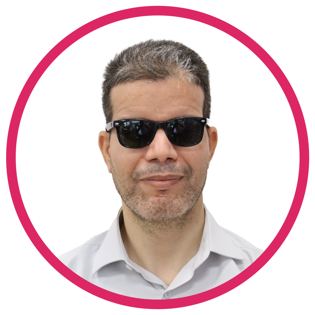 Portrait of Hamid A, a man with short, dark hair with some gray, wearing black sunglasses and a light-colored collared shirt. They are smiling slightly, set inside a circular frame with a pink border.