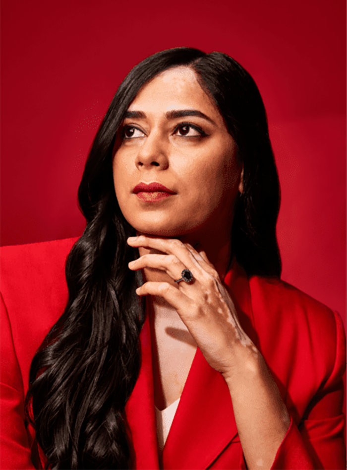 Alwar Pillai wearing a red blazer posing against a red backdrop. She is an Indian woman with brown skin and vitiligo and long, dark black hair.
