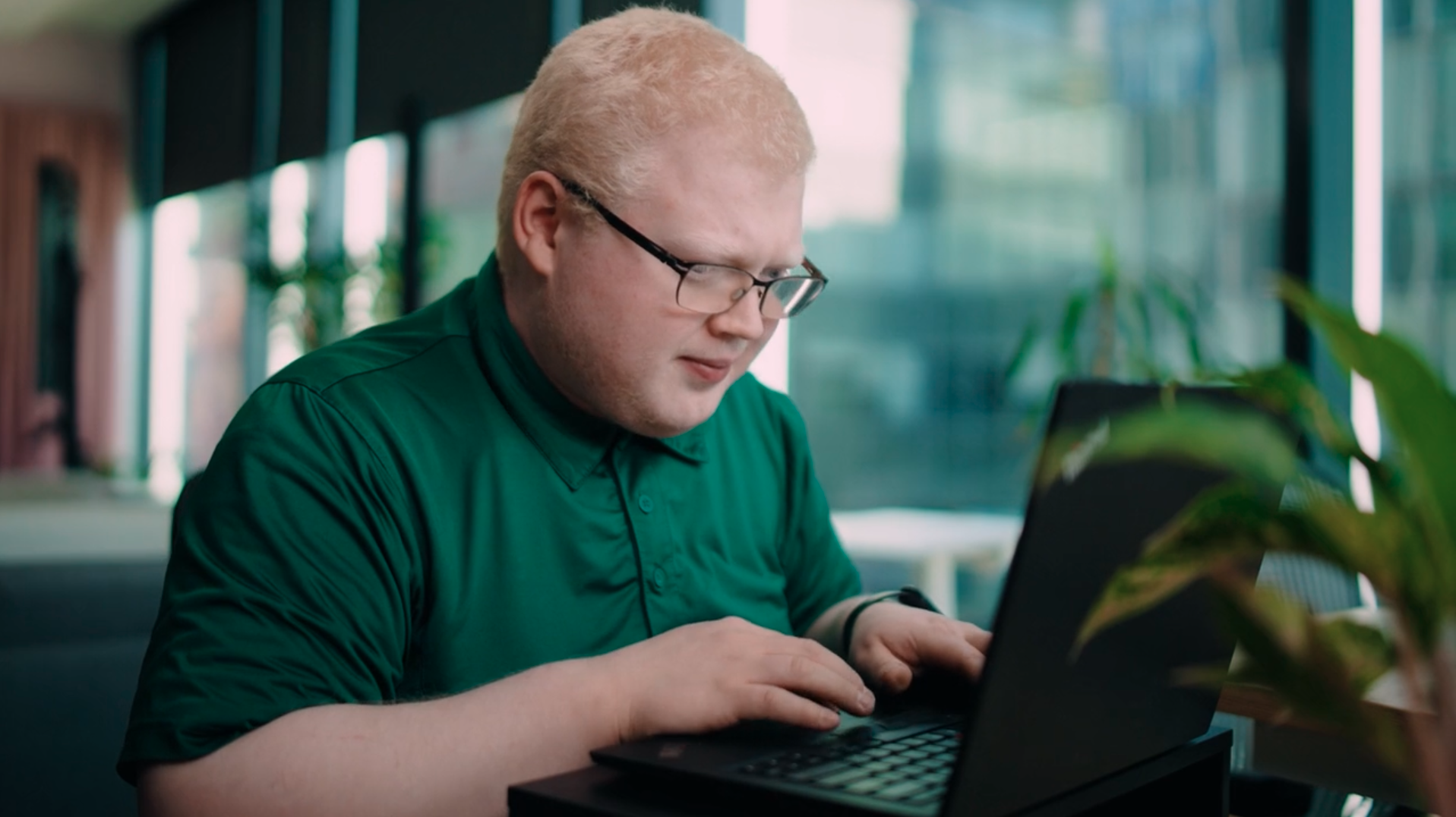 A man in an office wearing glasses and a green polo shirt sitting at a table and typing on a laptop. The individual is leaning in closely to the screen, with a focused expression.