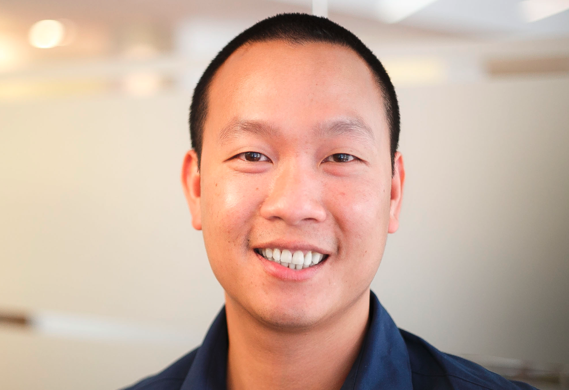 Toan Dinh, an asian man with short dark hair is smiling, wearing a dark blue shirt, shown against a blurred beige background.