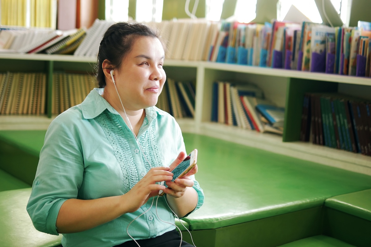 An Asian woman who is blind is sitting in a library, wearing earphones and holding a smartphone with both hands. She is smiling slightly. Bookshelves filled with books are visible in the background.