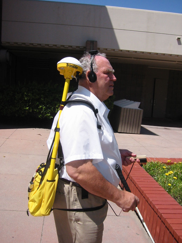 A blind man wearing a yellow backpack wth mounted GPS device. He is holding a white cane and wearing a headset, appearing to navigate an outdoor area with tiled pavement and a low brick planter.